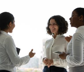 Team of happy female colleagues chatting near office window. Businesswomen standing together, talking, gesturing and smiling. Business networking concept