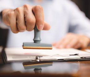 Close-up of a person's hand stamping with passed and approved stamp on certificate document public paper at desk, notary or business people work from home, isolated for coronavirus COVID-19 protection
