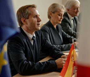 Caucasian middle aged man, Caucasian middle aged woman, and Caucasian senior man sitting at conference table participating in international meeting with European and Spanish flags visible